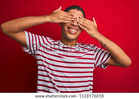 Young handsome arab man wearing striped t-shirt over isolated red background covering eyes with hands smiling cheerful and funny. Blind concept.