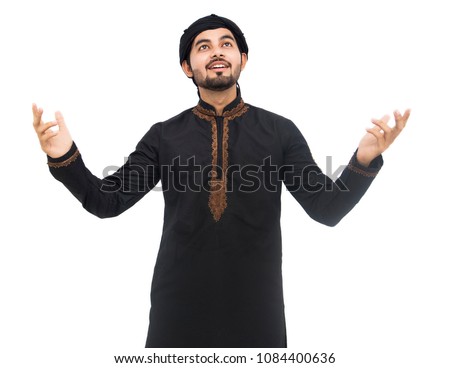 Young handsome Arabic Pakistani Indian Muslim boy man raising his hands with his eyes up and laughing while standing against white background and showing thumb on camera front in black kameez shalwar.