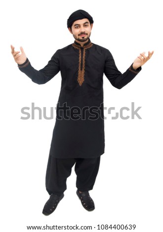 Young handsome Arabic Pakistani Indian Muslim boy man raising his hands with his eyes up and laughing while standing against white background and showing thumb on camera front in black kameez shalwar.