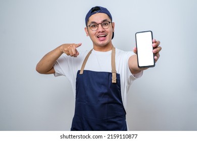 Young handsome asian barista man wearing apron pointing finger at mobile screen, showing app and smiling, standing over white background