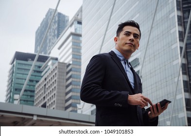 Young Handsome Asian Business Man Wearing Modern Black Suit Reading Information About Finance News With Mobile Smart Phone In Building City Background, Network Technology, Internet, Successful Concept