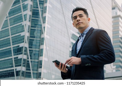 Young Handsome Asian Business Man Wearing Modern Black Suit Reading Information About Finance News With Mobile Smart Phone In Building City Background, Network Technology, Internet, Successful Concept