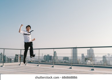 Young handsome Asian businessman jumping high, celebrate success winning pose on building rooftop. Work, job, or successful business concept. Cityscape background with copy space on sunny blue sky - Powered by Shutterstock