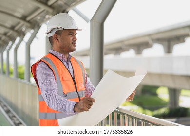 Young handsome asian civil engineer holding paperwork looking and planing standing at construction site. Worker wearing hard hat at highway concrete road site.