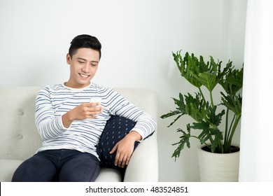 Young Handsome Asian Man Using Modern Mobile Smart Phone Sitting On A Couch At Home