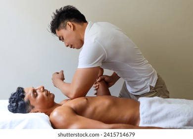 young handsome asian massage therapist perform massage on pectoralis for young muscular man who lying on the massage table in the supine position. White room background. - Powered by Shutterstock