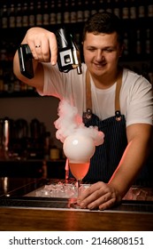 Young Handsome Barman Holds A Glass With Refreshing Orange Alcoholic Cocktil And Decorates It With Smoky Air Bubble. Shelves With Bottles On Background.