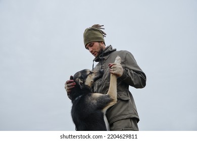 Young Handsome Bearded Caucasian Man Chooses Mongrel Dog In Animal Shelter. Minimalistic Portrait Outside Against Gray Sky. Male Owner Cuddles With Alaskan Husky Dog. Dog Put Its Paws On Man.