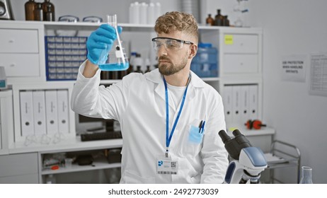 A young, handsome, bearded caucasian man in a lab coat examines a blue liquid in a flask inside a laboratory. - Powered by Shutterstock