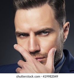 Young handsome bearded caucasian man with blue eyes and a hand near chin. Perfect skin and hairstyle. Wearing blue suit and watch.  Studio portrait on gradient black to grey background. Toned