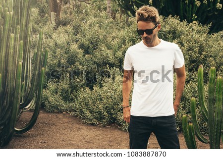 Young handsome bearded guy wearing a blank white t-shirt and sunglasses is standing in the garden background next to a big cactus plant. Horizontal mock up style