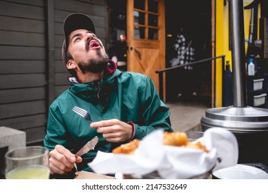 Young, Handsome, Bearded And Happy Man With Green Jacket And Trucker Cap Laughing While Eat Some Very Hot Deep-fried Snacks In The Outside In The Day