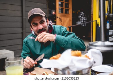 Young, Handsome, Bearded And Happy Man With Green Jacket And Trucker Cap Laughing Heartily While Eat Some Deep-fried Meat And Vegetable In The Outside In The Day