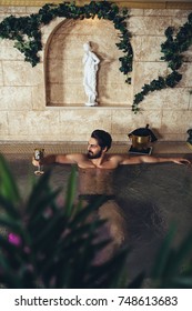 Young Handsome Bearded Man Enjoying And Relaxing In Spa Center. He Sitting In Jacuzzi Or Hot Tub And Drinking Wine.