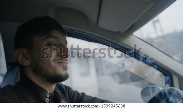 Young handsome bearded man in jacket smoking and\
driving in car, view from front passenger. Beautiful cityscape view\
out of the window