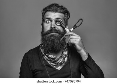 Young Handsome Bearded Man With Long Beard Moustache And Brunette Hair Holding Hairdresser Or Barber Scissors With Emotional Face In Studio On Grey Background