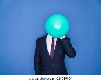 Young handsome bearded man in a suit blowing up a green balloon front view. Blue background.