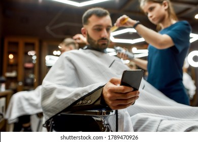 Young Handsome Bearded Man Visiting Barbershop, Sitting In Chair And Using His Smartphone While Professional Barber Girl Making Trendy Haircut For Him. Focus On Phone