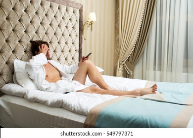 Young Handsome Bearded Man In White Terry Bathrobe Laying On Bed In Hotel Bedroom And Watching Tv With Remote Control