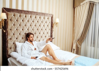 Young Handsome Bearded Man In White Terry Bathrobe Laying On Bed In Hotel Bedroom And Watching Tv With Remote Control