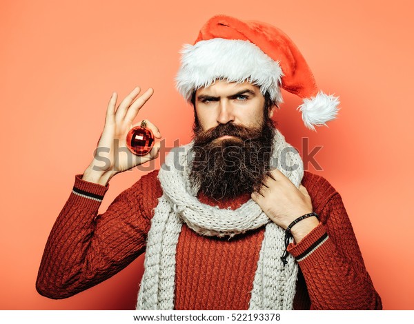 赤いセーターに長いひげ スカーフに新年の帽子をかぶった若いハンサムな髭を生やしたサンタクロースの男性が オレンジのスタジオの壁の背景に飾り付けたクリスマスまたはクリスマスボールを持つ の写真素材 今すぐ編集