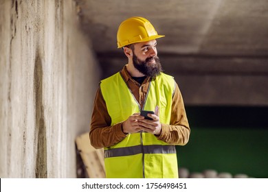 Young handsome bearded worker standing in front of concrete wall and using smart phone.