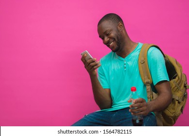 Young handsome black man carrying a backpack and smiling while pressing his phone sitting down with a bottle of drink