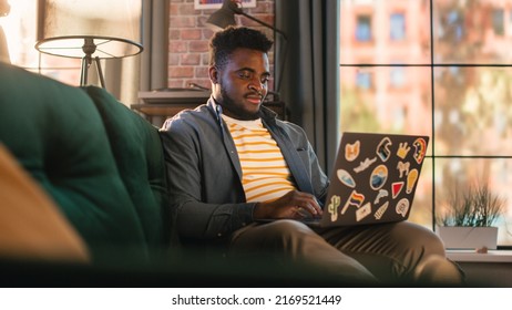 Young Handsome Black Man Working from Home on Laptop Computer in Sunny Stylish Loft Apartment. Creative Male Checking Social Media and Browsing Internet on a Sofa. Urban City View from Big Window.