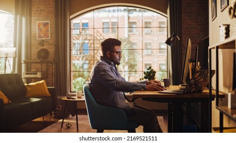 Young Handsome Black Man Working from Home on Desktop Computer in Sunny Stylish Loft Apartment. Creative Male Checking Social Media, Browsing Internet. Urban City View from Big Window.