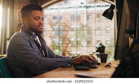 Young Handsome Black Man Working from Home on Desktop Computer in Sunny Stylish Loft Apartment. Creative Male Checking Social Media, Browsing Internet. Urban City View from Big Window.