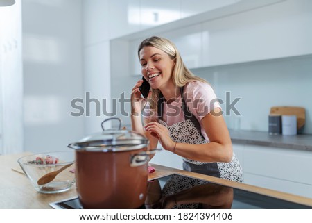 Young handsome blond lazy housewife standing in kitchen, talking on the phone while pot with lunch is on the stove.