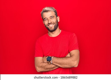 Young Handsome Blond Man Wearing Casual Red T-shirt Standing Over Isolated Red Background Happy Face Smiling With Crossed Arms Looking At The Camera. Positive Person.