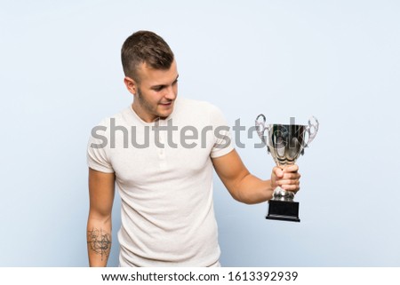 Young handsome blonde man over isolated blue background holding a trophy