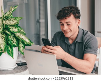 Young handsome boy while using his smartphone typing message sitting on outdoor table. A laptop is open in front of him, suggesting he might be working or studying remotely