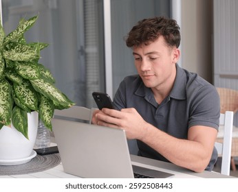 Young handsome boy while using his smartphone typing message sitting on outdoor table. A laptop is open in front of him, suggesting he might be working or studying remotely