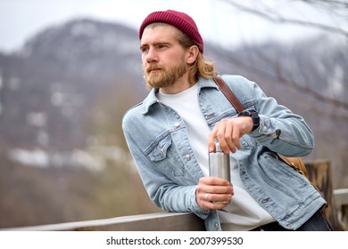 Young Handsome Brutal Bearded Man Traveling In The Wild Nature With Thermos, Autumn Or Spring Style, Traveler In Casual Wear Have Rest, Looking At Side On Landscapes, Adventure, Free Spirit