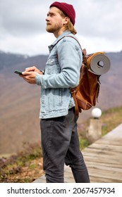 Young Handsome Brutal Bearded Man Traveling In The Wild Nature Using Mobile Phone, With Leather Brown Backpack, Autumn Style, Traveler, Adventure, Free Spirit. Copy Space. Side View