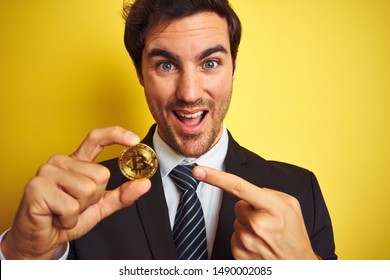 Young handsome businessman holding bitcoin standing over isolated yellow background very happy pointing with hand and finger Stock fotografie