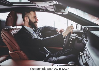 Young Handsome Businessman Is Sitting In Luxury Car. Serious Bearded Man In Suit Is Driving.