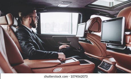 Young Handsome Businessman Is Sitting In Luxury Car. Serious Bearded Man In Suit And Glasses