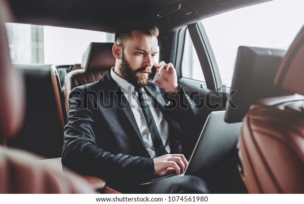 Young handsome businessman is sitting in\
luxury car. Serious bearded man in suit is working with laptop and\
talking on smart phone while being in\
trip.