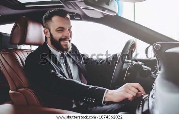 Young handsome businessman is sitting\
in luxury car. Serious bearded man in suit is\
driving.