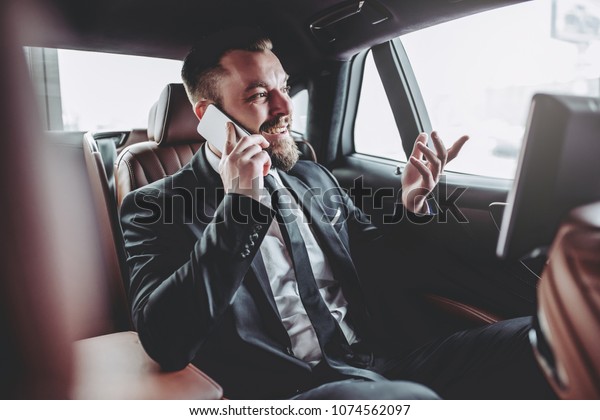Young handsome businessman is sitting in\
luxury car. Serious bearded man in suit is working with laptop and\
talking on smart phone while being in\
trip.