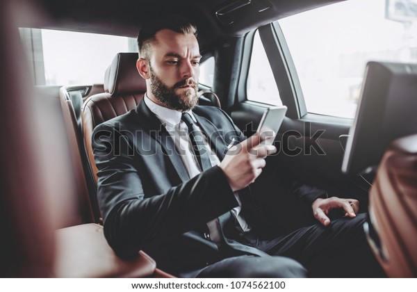 Young handsome businessman is sitting in luxury\
car. Serious bearded man in suit is working with smart phone while\
being in trip.