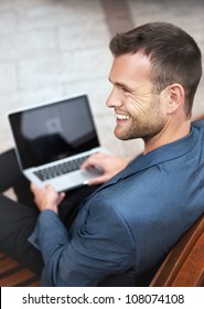 Young handsome businessman sitting on a wooden bench in the city with a laptop and smiling. From above