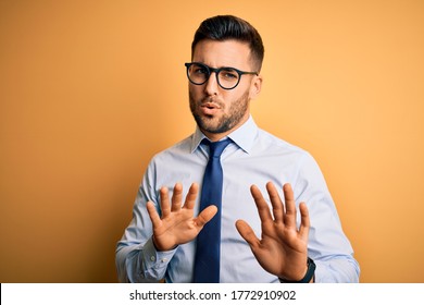 Young handsome businessman wearing tie and glasses standing over yellow background Moving away hands palms showing refusal and denial with afraid and disgusting expression. Stop and forbidden. - Powered by Shutterstock