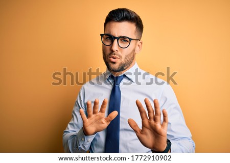 Young handsome businessman wearing tie and glasses standing over yellow background Moving away hands palms showing refusal and denial with afraid and disgusting expression. Stop and forbidden.