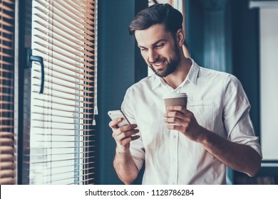 Young And Handsome Businessman In White Shirt With Cup Of Coffe And Mobile Phone In Hands Near Windows In Modern Office. Happy Office Worker At Work. Business Concept. Guy With Gadget.