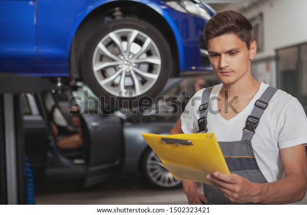 Young handsome car mechanic reading documents on\
his clipboard while repairing a car at the garage. Car service,\
maintenance concept