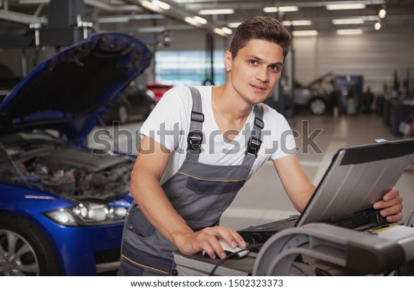 Young handsome car mechanic smiling\
to the camera, using laptop for computer diagnostics of a broken\
vehicle. Cheerful car technician working at the\
garage